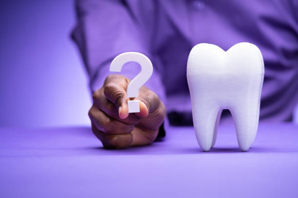 A person in a purple shirt holding up a white question mark beside a large model of a tooth in front of a purple background.