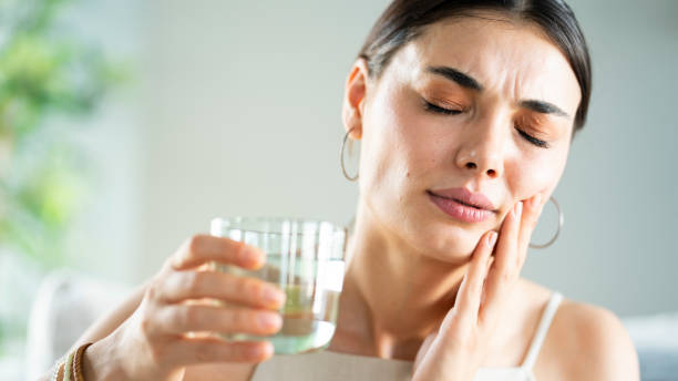 Woman with tooth pain touches her jaw and holds a glass of water in her other hand